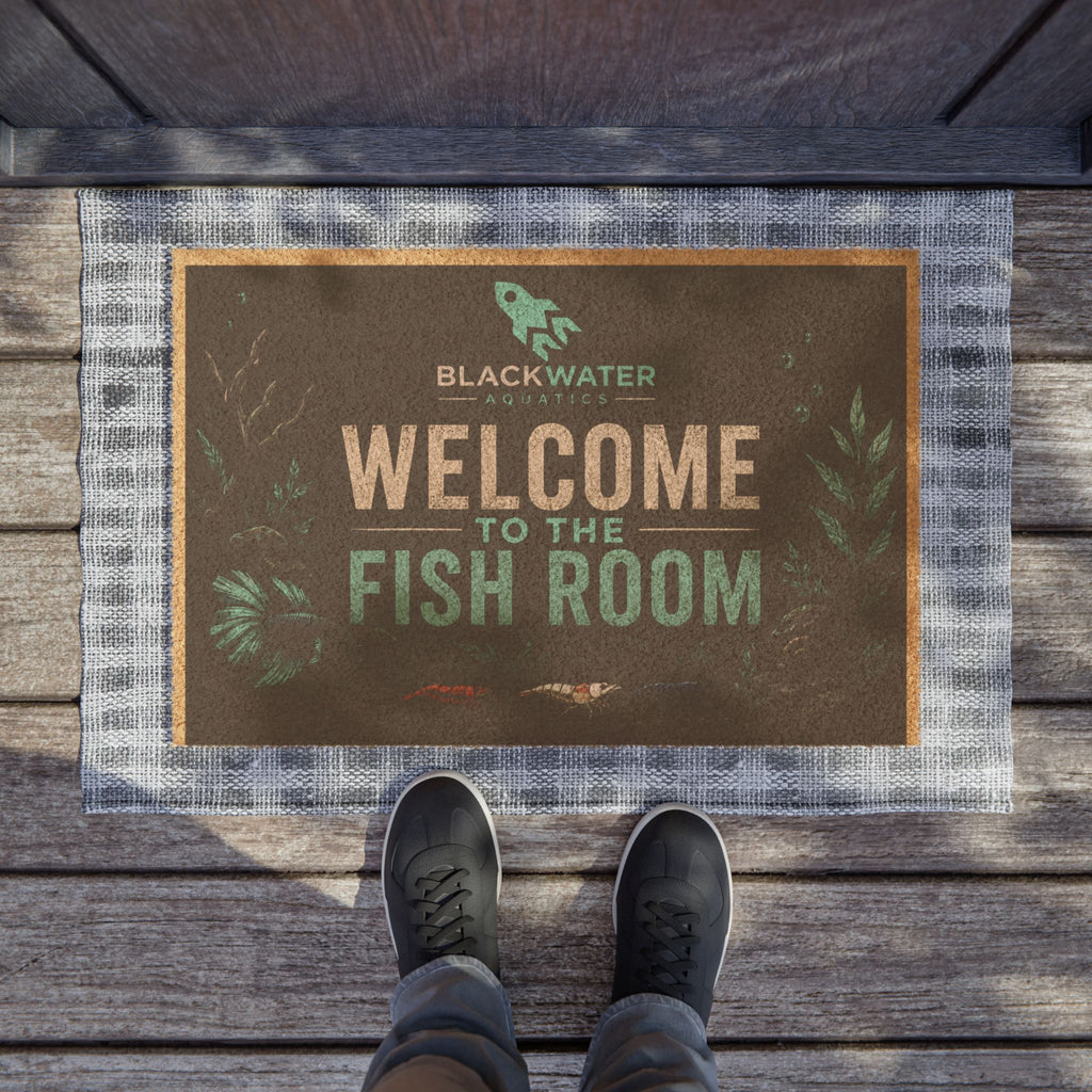 A rectangular doormat with a rustic brown center panel and a gray plaid border, placed on a wooden deck. The mat reads “WELCOME TO THE FISH ROOM” with “BLACKWATER ADVENTURES” above it.