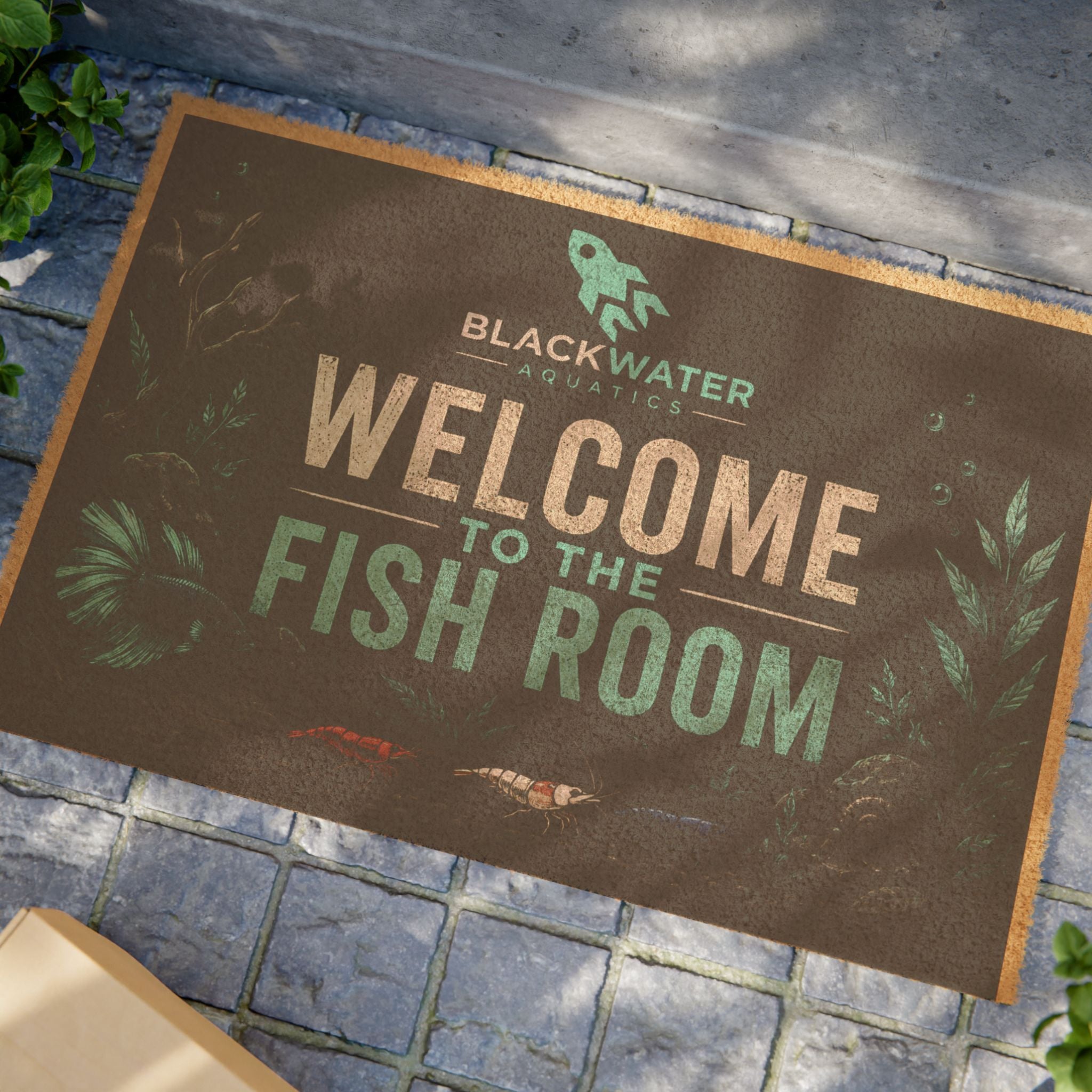 A rectangular printed mat or sign laid on gray stone tiles, featuring the text 'BLACKWATER AQUATICS WELCOME TO THE FISH ROOM' with fish and aquatic plant illustrations. No person or animal is present, but this is not a clean isolated studio product shot because a real outdoor/indoor stone surface is visible, so Lifestyle fits better than Product/Packaging.