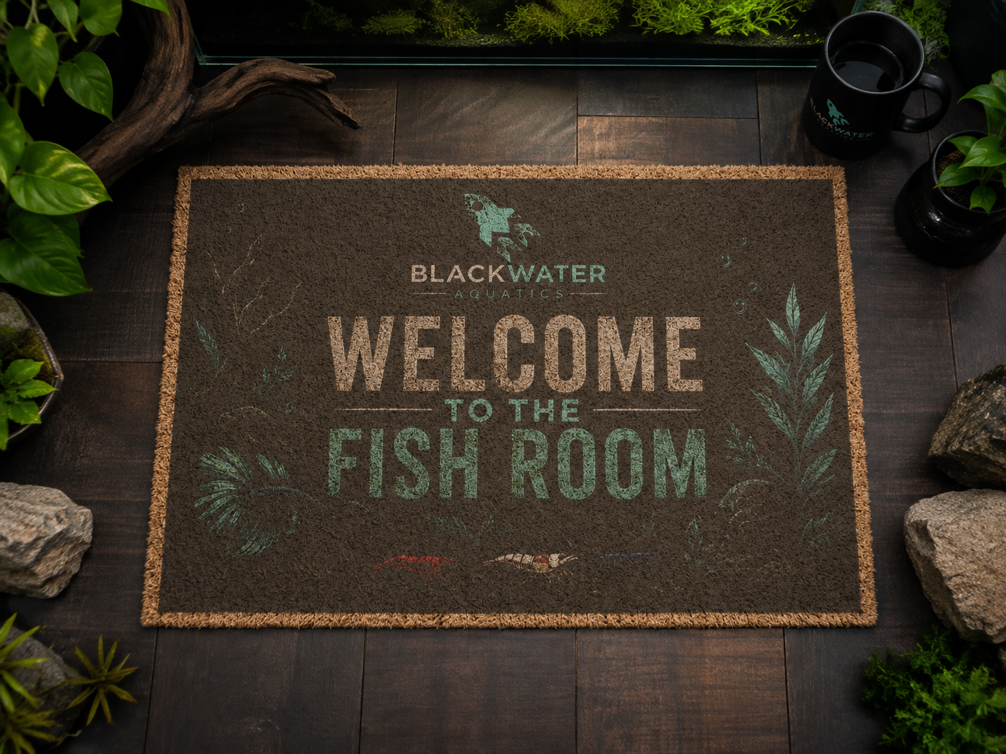 A rectangular coir doormat with the text 'BLACKWATER EXPERIENCE WELCOME TO THE FISH ROOM' lies on a dark wood floor, surrounded by plants, rocks, and a black mug.
