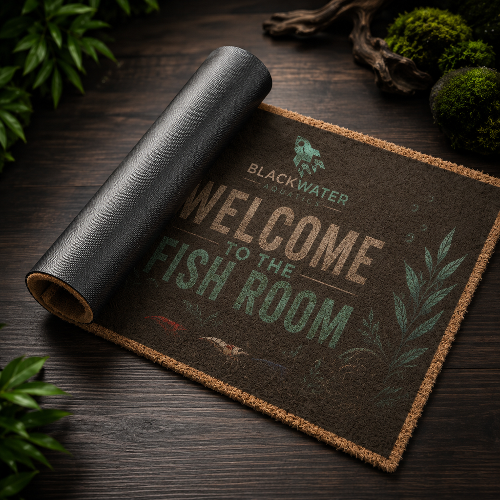 A rolled black coir-style doormat with printed text reading “BLACKWATER FISHING” and “WELCOME TO THE FISH ROOM,” photographed on a dark wooden surface with decorative moss and driftwood around it.
